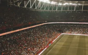 Estadio de futbol iluminado durante un partido nocturno con las gradas llenas