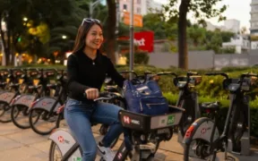 Mujer sonríe sobre una bicicleta compartida junto a una cicloestación al atardecer