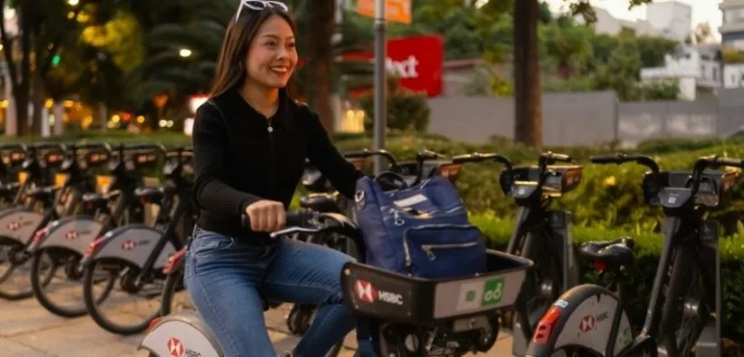 Mujer sonríe sobre una bicicleta compartida junto a una cicloestación al atardecer