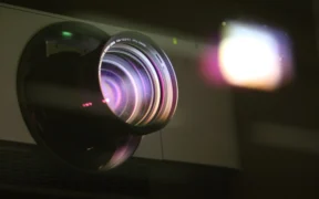 Detalle de la lente iluminada de un proyector; fotografía contextual de Ann H