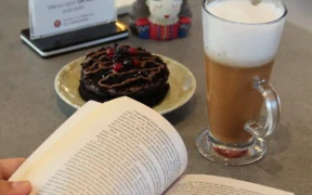 Manos de una persona con un libro abierto junto a café y postre sobre una mesa.