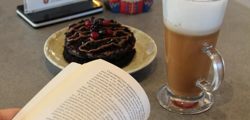 Manos de una persona con un libro abierto junto a café y postre sobre una mesa.